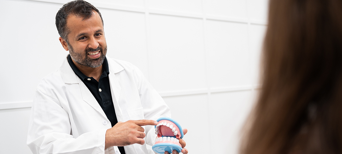 Dr. Dhillon, smiles while holding a dental model and pointing to the teeth during a patient consultation in a brightly lit dental office.